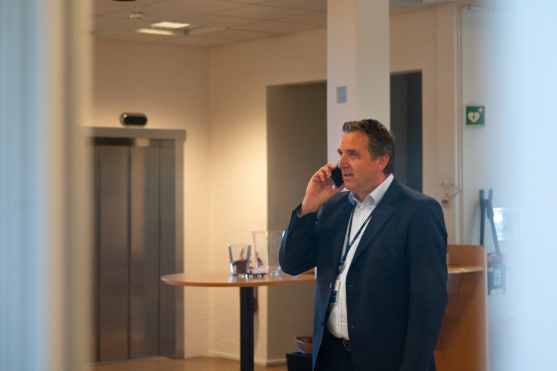 Adm. dir. Arne Martin Laukvik står med telefonen smilende opp på en skjerm. Bilde tatt gjennom vinduet.